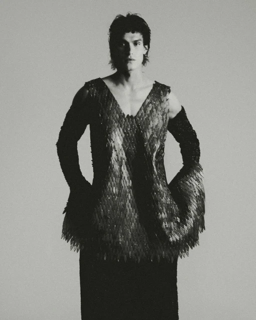 Monochrome portrait of a male model wearing a unique v-neck top with scale-like embellishments and fitted black arm sleeves, against a light grey backdrop.