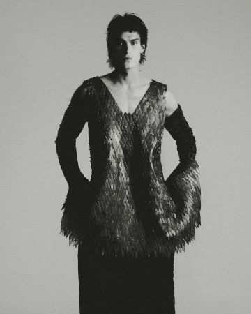 Monochrome portrait of a male model wearing a unique v-neck top with scale-like embellishments and fitted black arm sleeves, against a light grey backdrop.