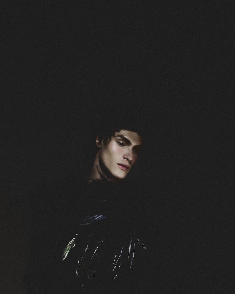 Portrait of a young adult male with dark hair and pale skin in dark lighting, looking down, wearing a dark textured top.