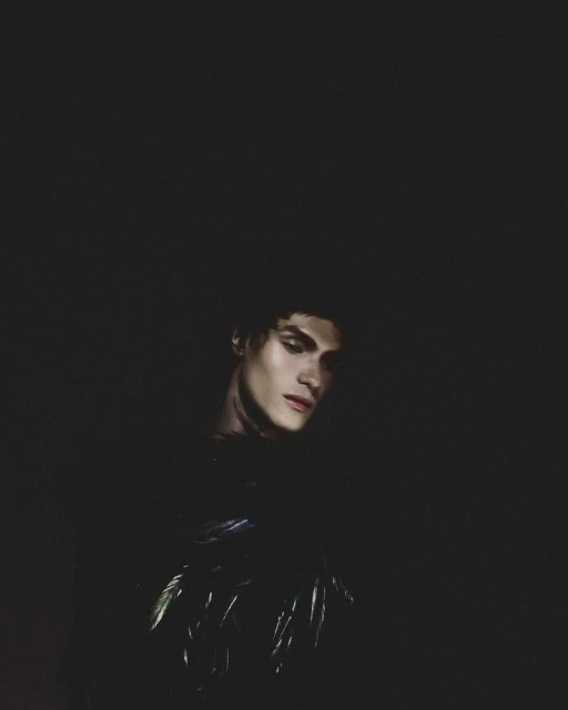 Portrait of a young adult male with dark hair and pale skin in dark lighting, looking down, wearing a dark textured top.