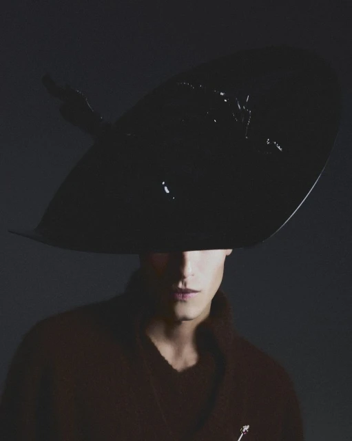 Elegant portrait of a person wearing a large black hat and a maroon coat, set against a dark background in a studio environment.