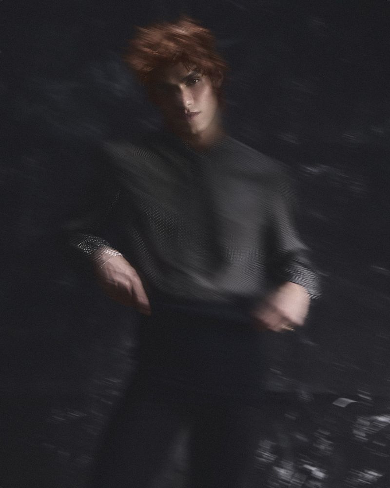 A young adult man with red hair and a black long-sleeve shirt poses in a dimly lit, blurred setting, creating a somber and artistic portrait.