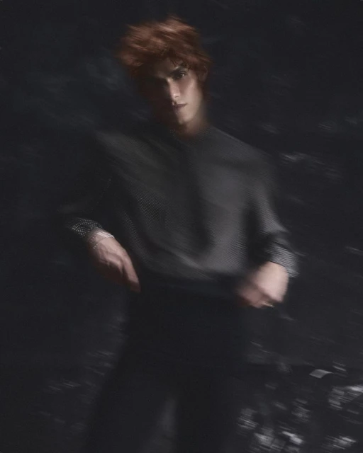 A young adult man with red hair and a black long-sleeve shirt poses in a dimly lit, blurred setting, creating a somber and artistic portrait.
