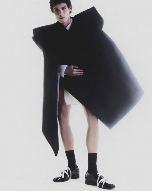 A person wears an oversized, angular black jacket with a white shirt, short, and black socks with black shoes in a studio shot.