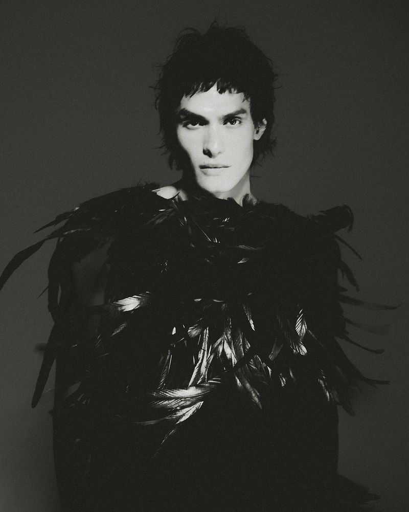 Monochrome portrait of a young adult male with dark wavy hair, wearing a black top adorned with feathers, against a dark gray backdrop.