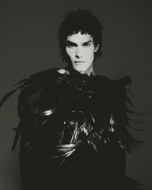 Monochrome portrait of a young adult male with dark wavy hair, wearing a black top adorned with feathers, against a dark gray backdrop.