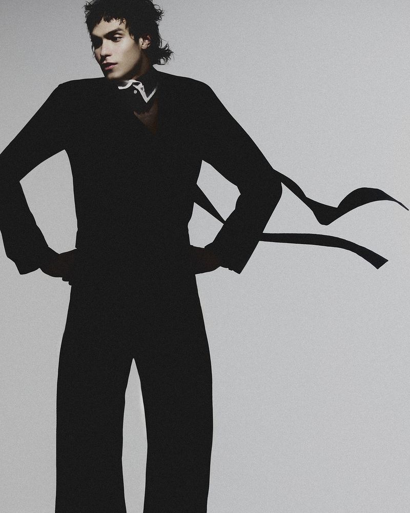 Young adult poses indoors in black suit with flowing ribbons. Expressive androgynous person stands against gray backdrop in fashion shot.