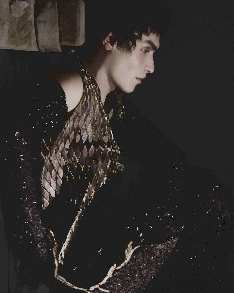 Portrait of a male fashion model wearing a gold scale vest and a black sequin shirt against a dark backdrop in a photo studio.