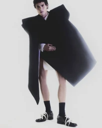 Model wearing an oversized dark geometric coat, white socks, and black lace-up shoes against a plain white studio background.