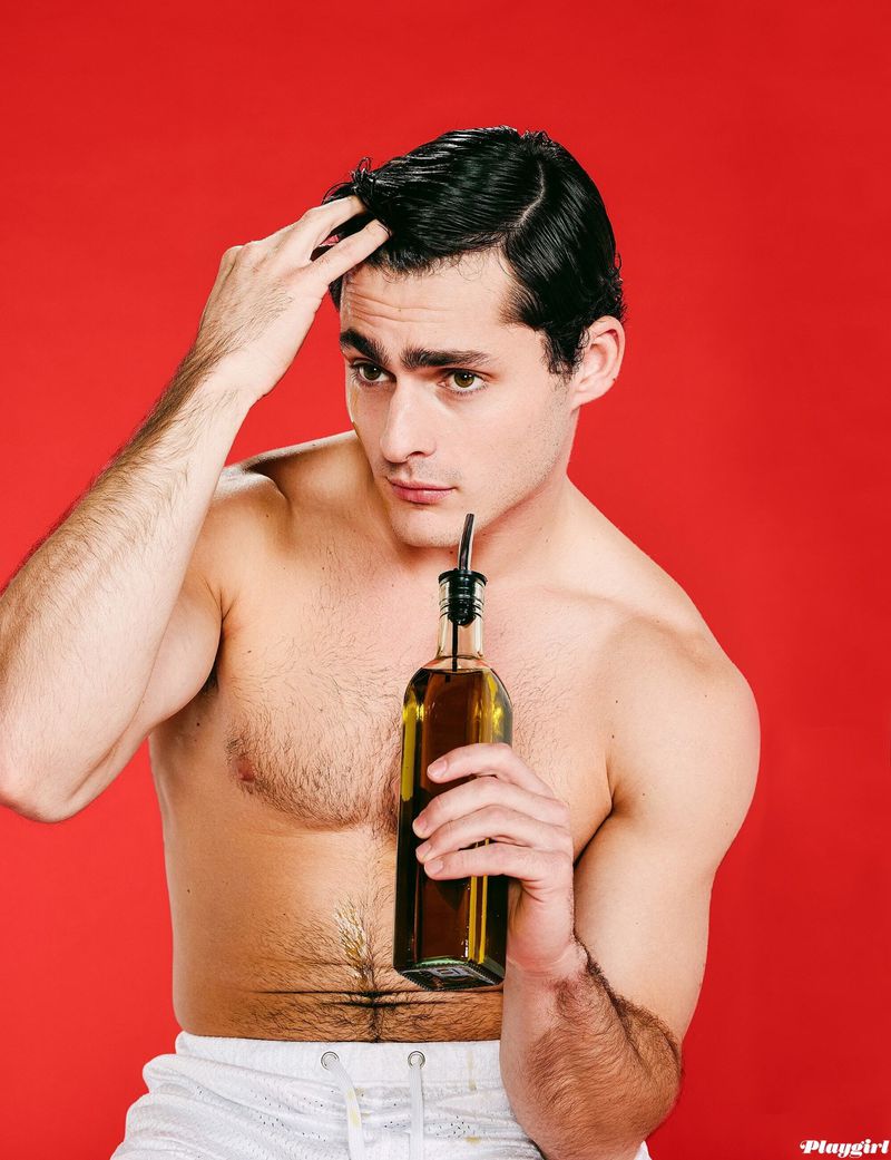 A man with a bottle of olive oil is pictured against a red background. He is shirtless, wearing shorts and styling his hair.