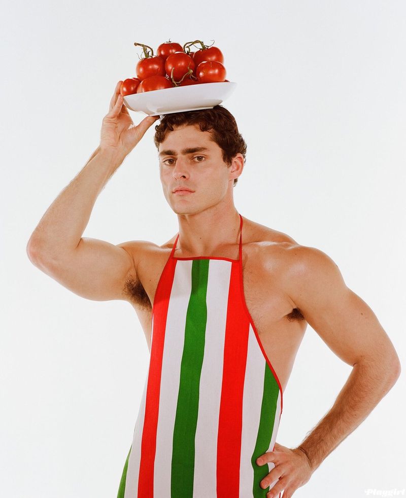 Man balances plate of tomatoes on head, wearing Italian-striped apron, posing in a studio with neutral expression.
