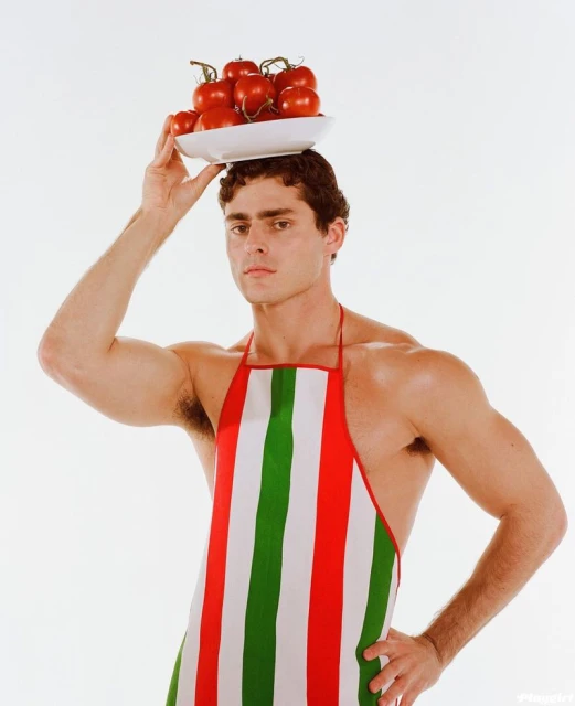 Man balances plate of tomatoes on head, wearing Italian-striped apron, posing in a studio with neutral expression.