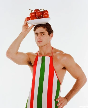 Man balances plate of tomatoes on head, wearing Italian-striped apron, posing in a studio with neutral expression.