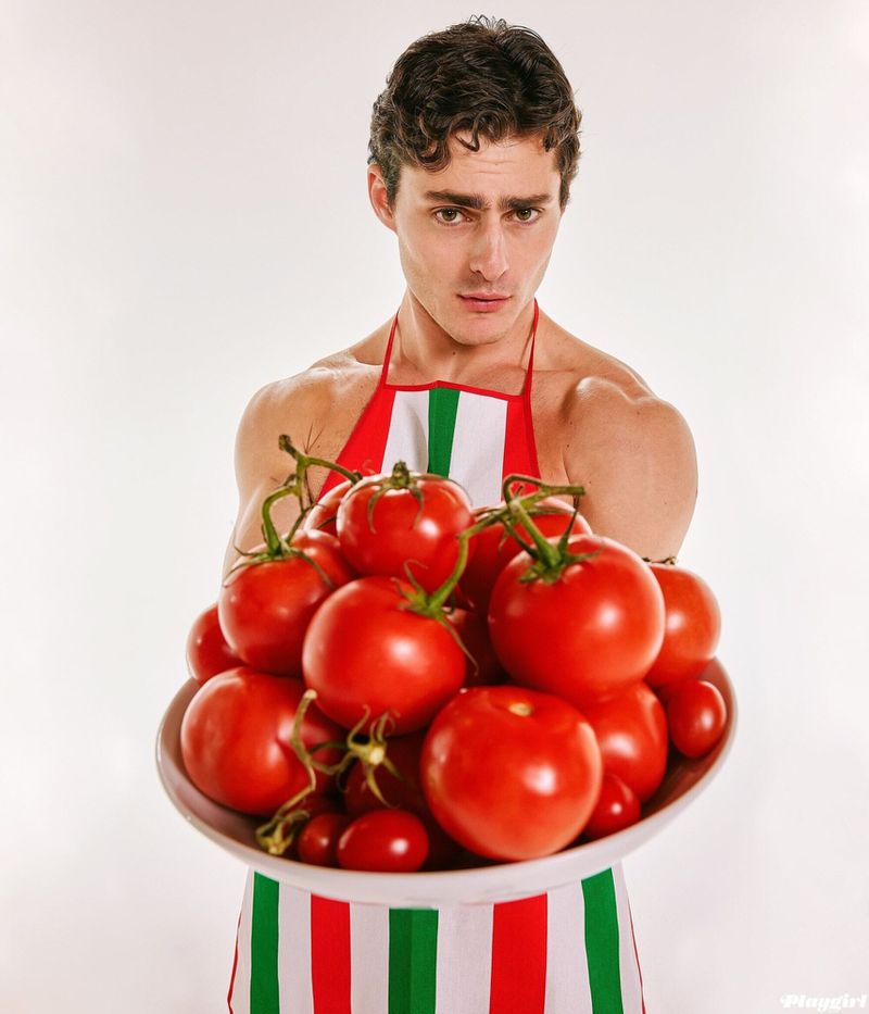 Attractive male model holds a bowl of red tomatoes while wearing a red, white, and green apron in a brightly lit photo studio.