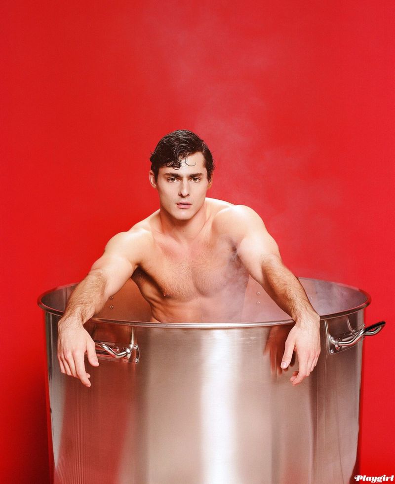 Shirtless adult male model sitting in a metal stockpot with a red backdrop and steam rising around him in a photo studio.