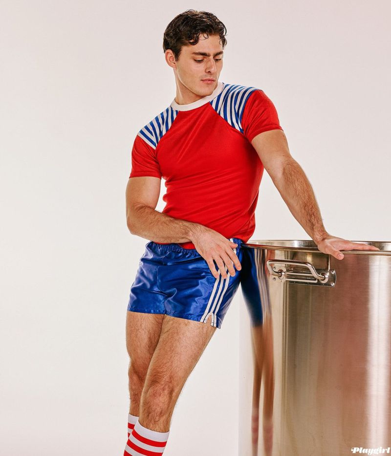 Athletic man in red and blue sportswear leans against a large stainless-steel container against a white backdrop in a studio setting.