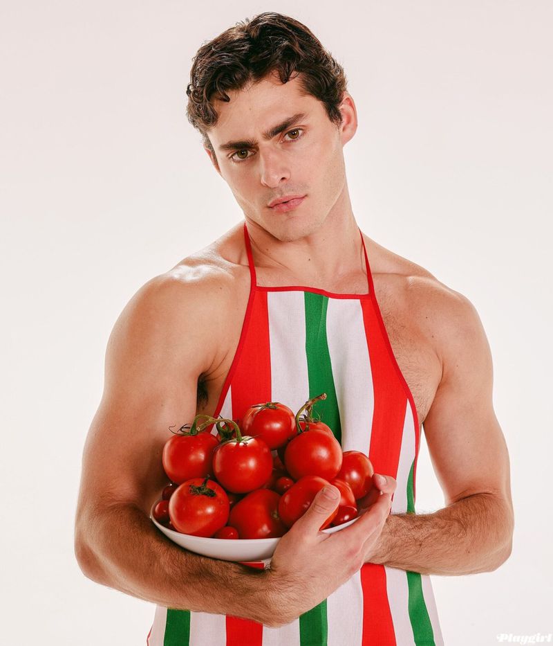 Man in striped apron holding bowl of fresh tomatoes against white backdrop, portrait orientation, with food and cooking theme.