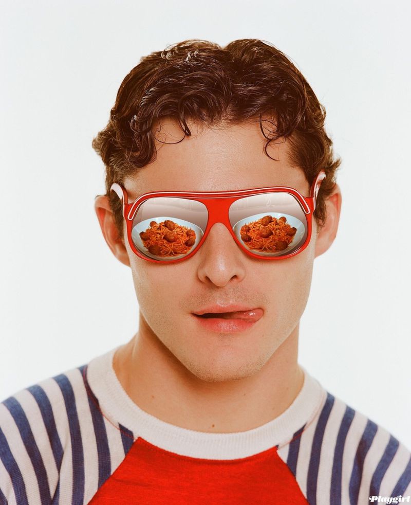 Close-up of a young man with dark hair, wearing red glasses reflecting two bowls of spaghetti, against a clean white backdrop.