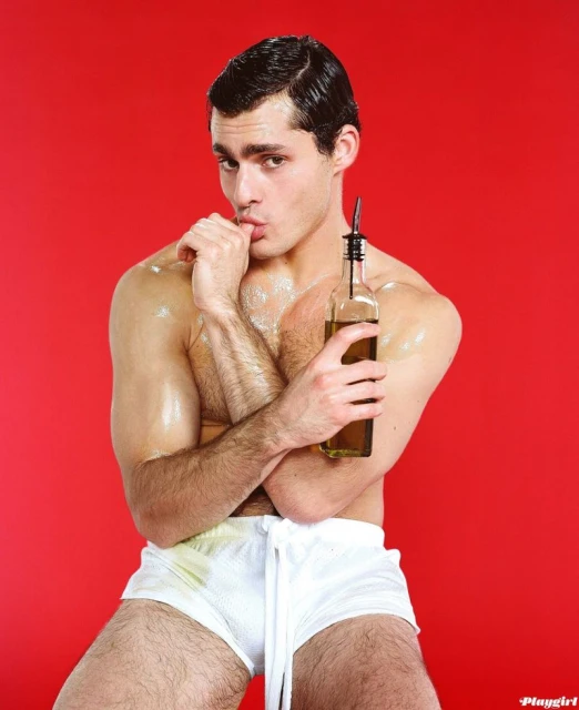Nude man in white shorts poses holding a bottle with a metal pour spout against a vibrant red background, studio lit.