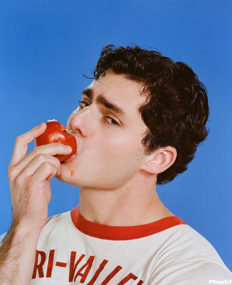 Portrait of a young adult male with dark curly hair eating a tomato against a bright blue background wearing a white shirt with red collar.