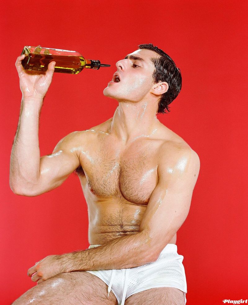 Shirtless man pouring a glass bottle into his mouth against a solid red studio background