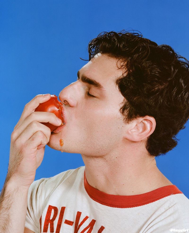 Close-up of a man with dark hair eating a red fruit, set against a vivid blue backdrop, wearing a casual t-shirt. 
