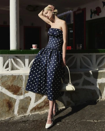 A fair-skinned woman poses in a navy blue polka dot dress and white heels, holding a white purse in front of a building.