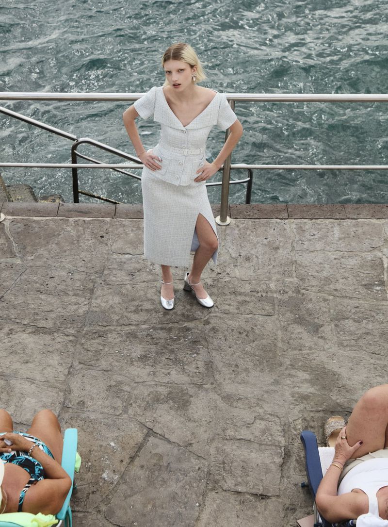 A woman in a matching top and skirt stands on a stone platform near the ocean, while other people recline on lounge chairs.
