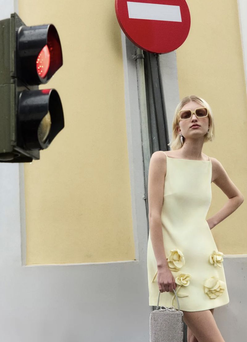 Woman in yellow dress with floral details posing near traffic light and street sign, holding a silver handbag, fashion outdoor shoot.