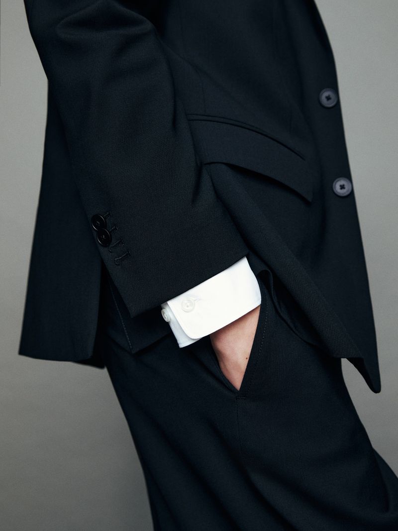 A person in a dark business suit with a white shirt, standing indoors against a gray background, showing the sleeve buttons and pocket.