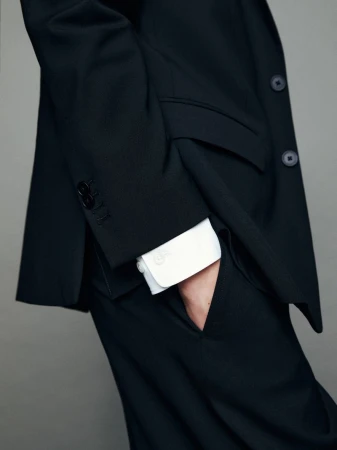 A person in a dark business suit with a white shirt, standing indoors against a gray background, showing the sleeve buttons and pocket.