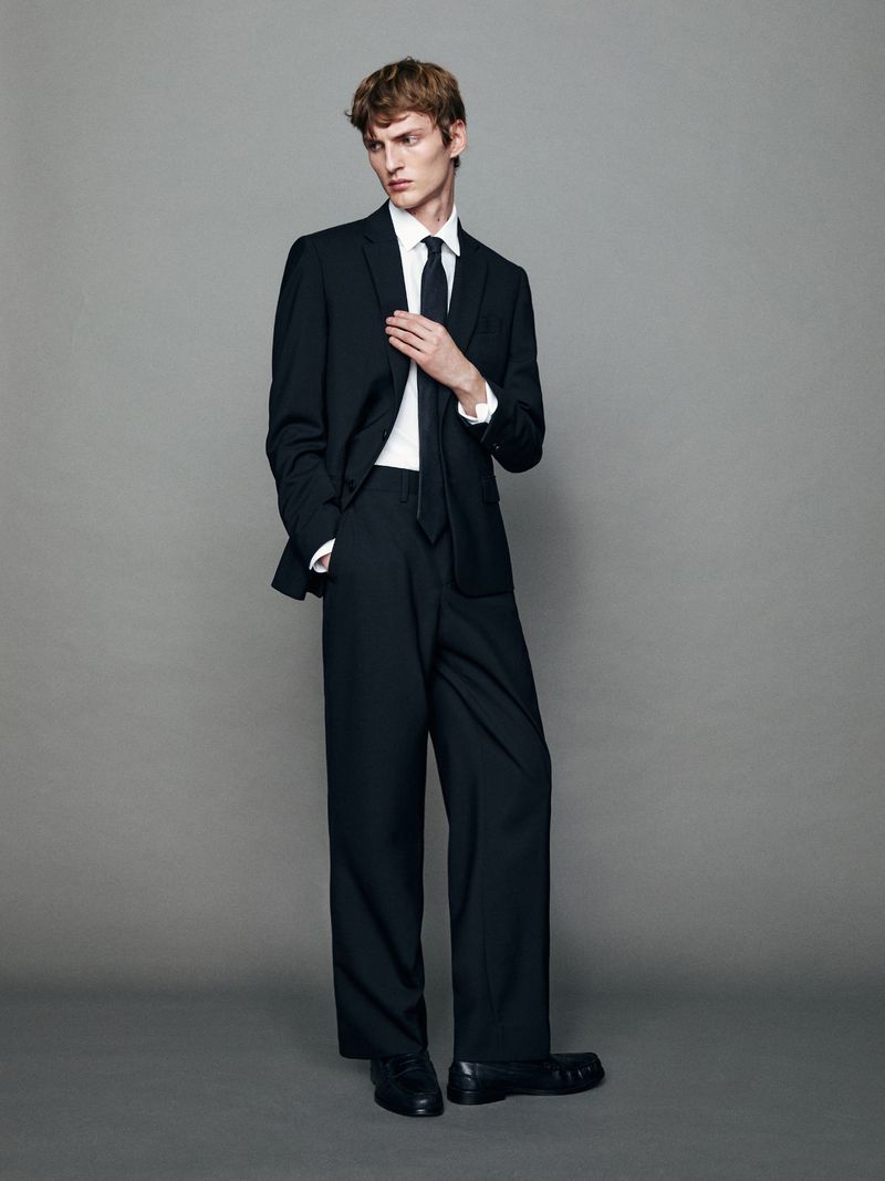 Model in a black suit with a white shirt and black tie standing against a neutral grey studio background for Massimo Dutti.