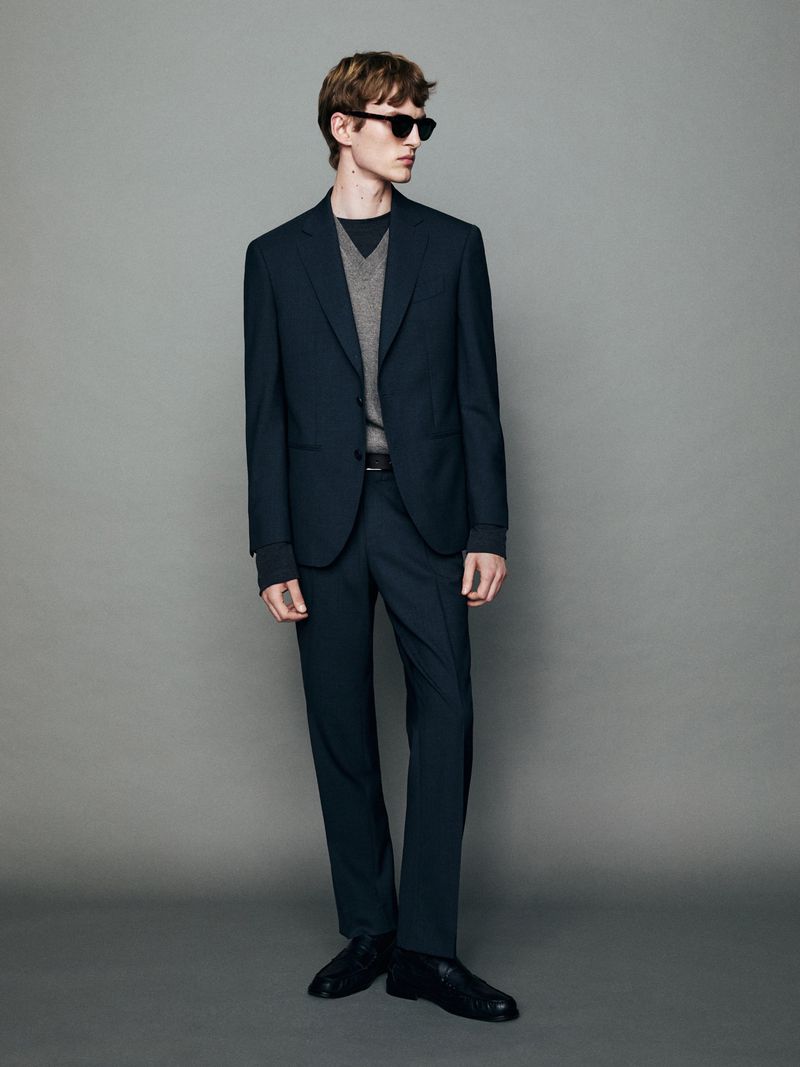 Model wearing dark suit jacket and trousers with sunglasses against a grey seamless studio background.