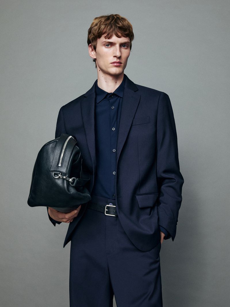 A young adult male poses in a studio wearing a navy suit, holding a black leather duffel bag in a fashion style against a grey backdrop.