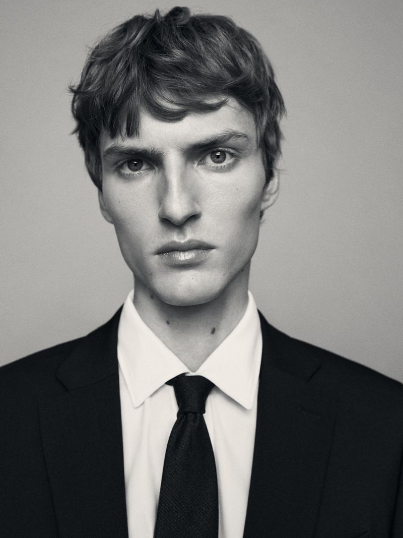 Portrait of a man wearing a black suit jacket, white shirt, and black tie against a grey background.