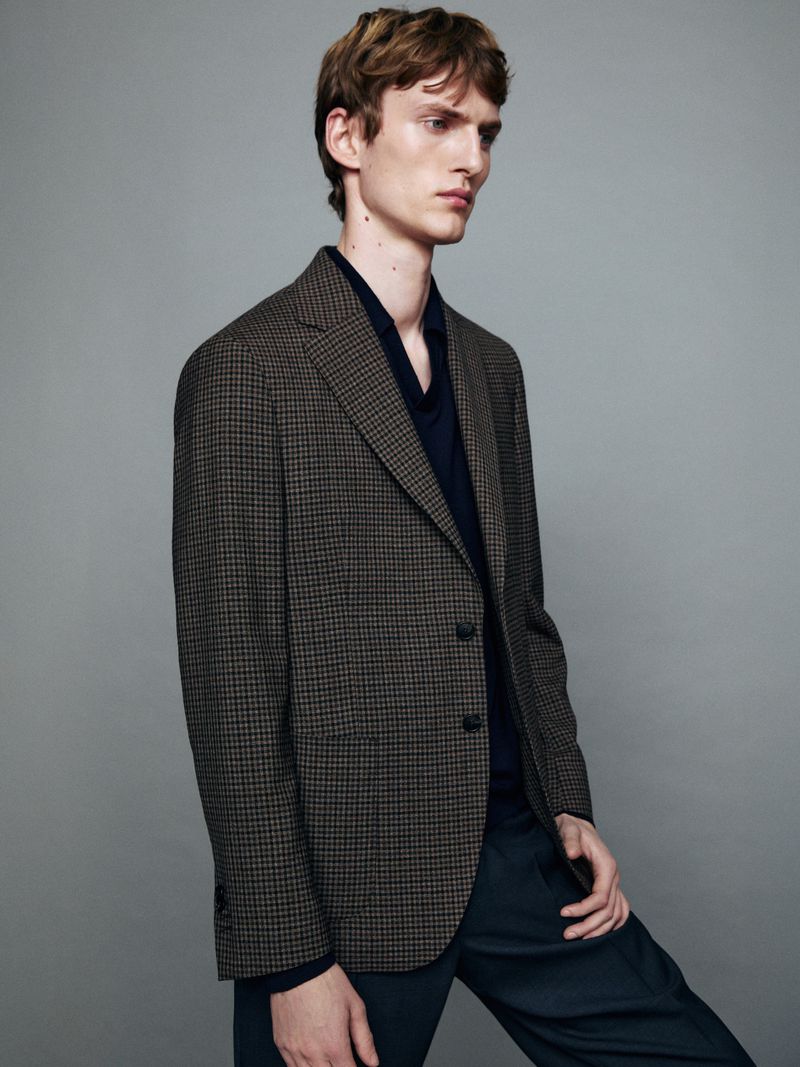 A fair-skinned adult male poses indoors wearing a brown checked blazer, unbuttoned navy shirt, and dark trousers against a gray backdrop.