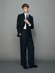 Model in a black suit with a white shirt and black tie standing against a neutral grey studio background for Massimo Dutti.