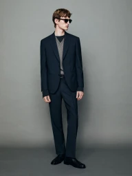 Model wearing dark suit jacket and trousers with sunglasses against a grey seamless studio background.