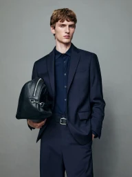 Male model in a navy suit holding a black leather duffel bag against a solid grey background