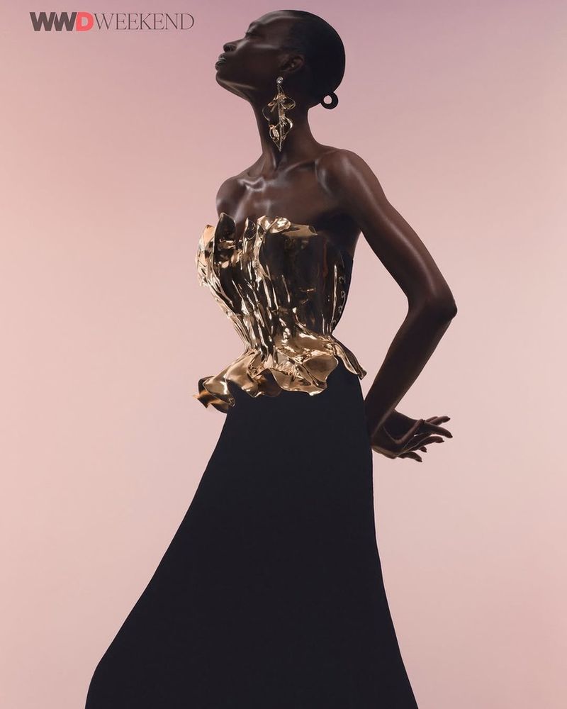 Editorial fashion shot of a slender Black model in a strapless gold corset and black dress against a pale pink background wearing statement earrings.