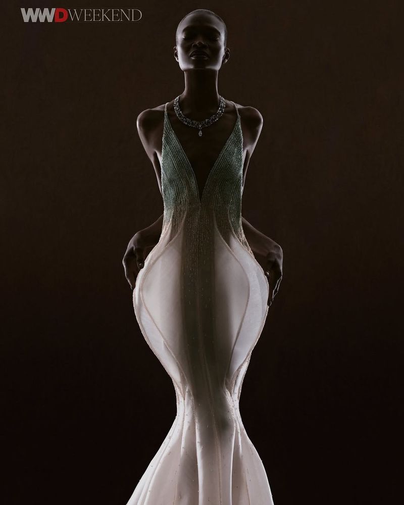 An adult female fashion model is illuminated in a full shot, wearing a white beaded gown and necklace against a dark backdrop.