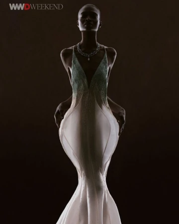 An adult female fashion model is illuminated in a full shot, wearing a white beaded gown and necklace against a dark backdrop.