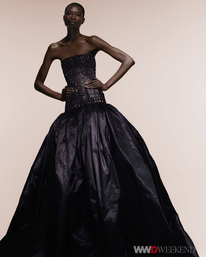 Fashion editorial shot of Black female model in strapless dark formal gown with hands on hips, posing against a neutral studio backdrop.