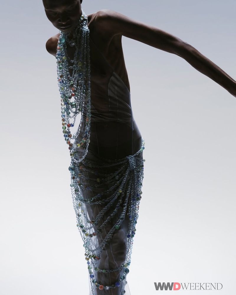 Woman in sheer draped garment and beaded necklaces stands in a brightly lit studio. Fashion editorial shot with soft lighting.