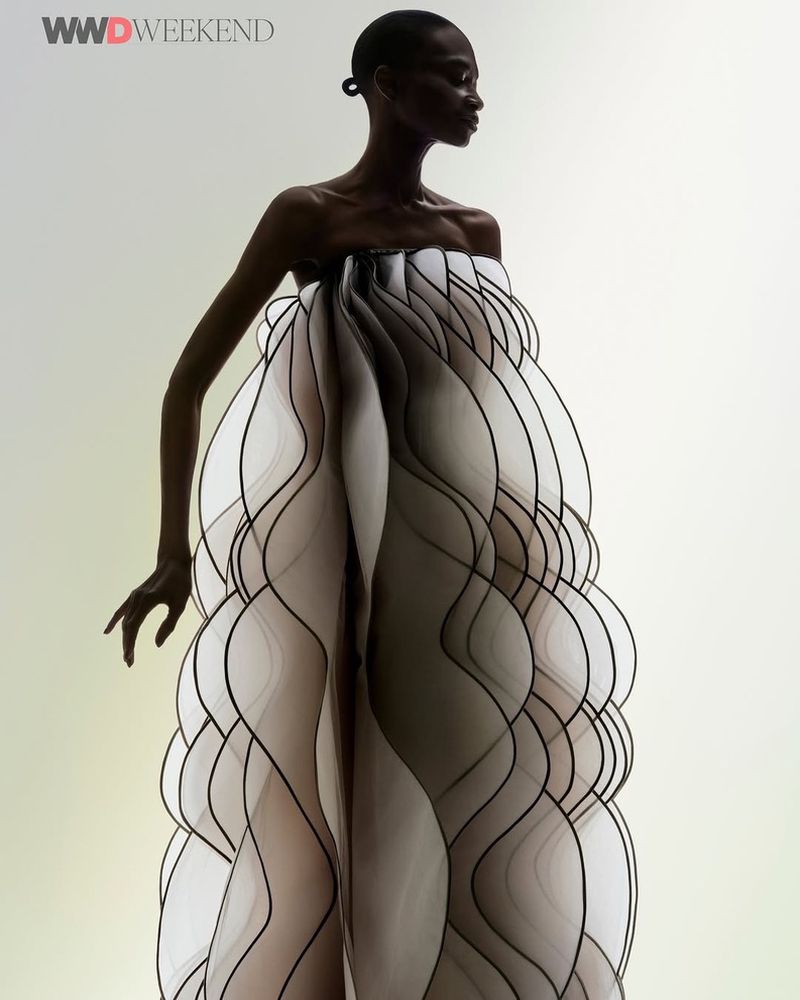 Striking editorial fashion shot: a slender Black woman stands in a voluminous, sculptural white dress with black accents against a pale backdrop.