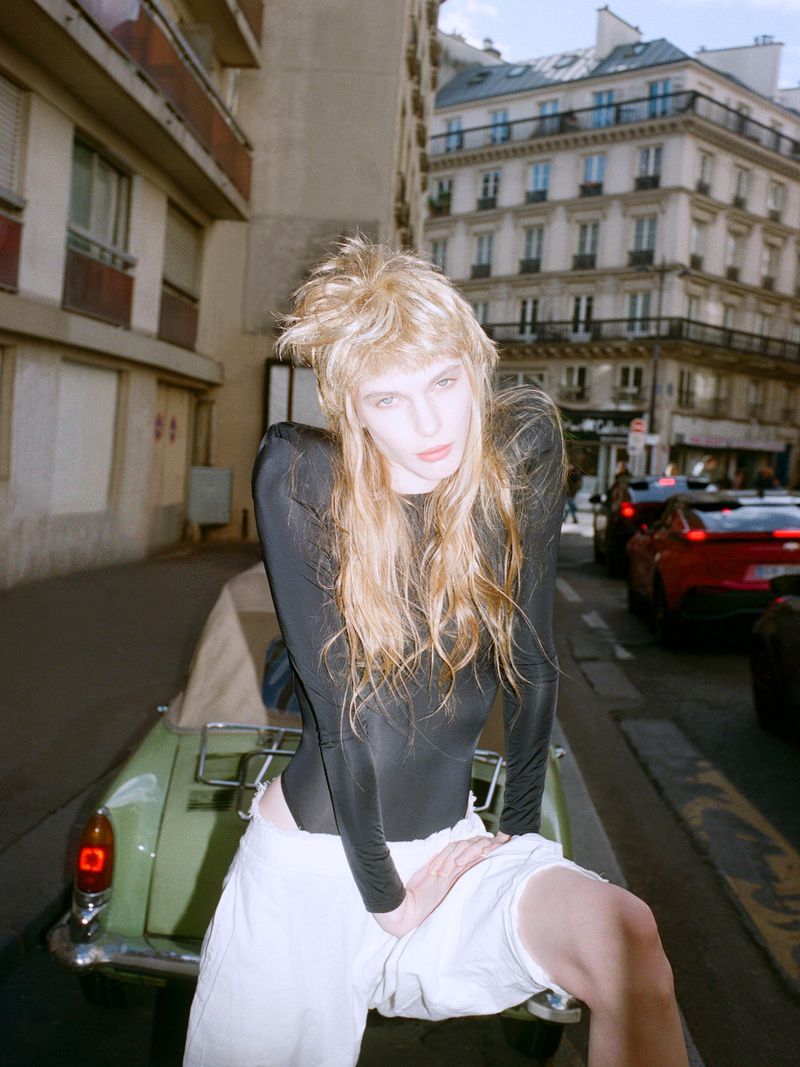 A blonde model poses on a vintage green car in the city, wearing a black bodysuit and white shorts against an urban backdrop.