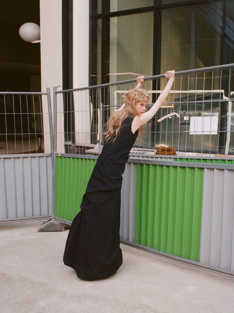 Woman in a black dress stands outdoors holding onto a metal and green barrier, with a building in the background, captured in natural light.
