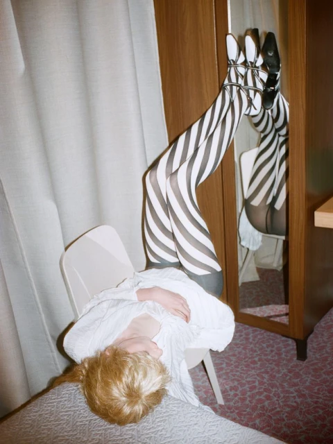 Woman in striped tights lying on a bed with her legs up against a mirror. The image features a playful and unconventional pose indoors.