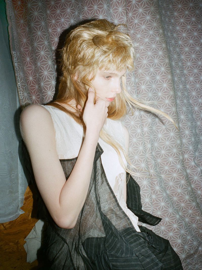 A young woman with blond hair is looking down and holding her hand up to her face, wearing a white and black sleeveless top.
