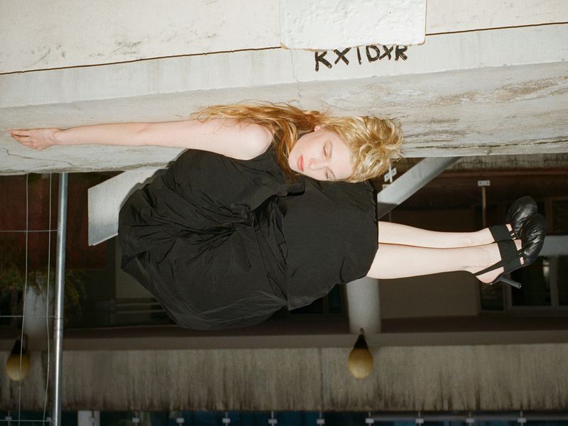 A young woman with red hair and wearing a black dress poses in an unusual pose and wearing high heels in a low light setting.